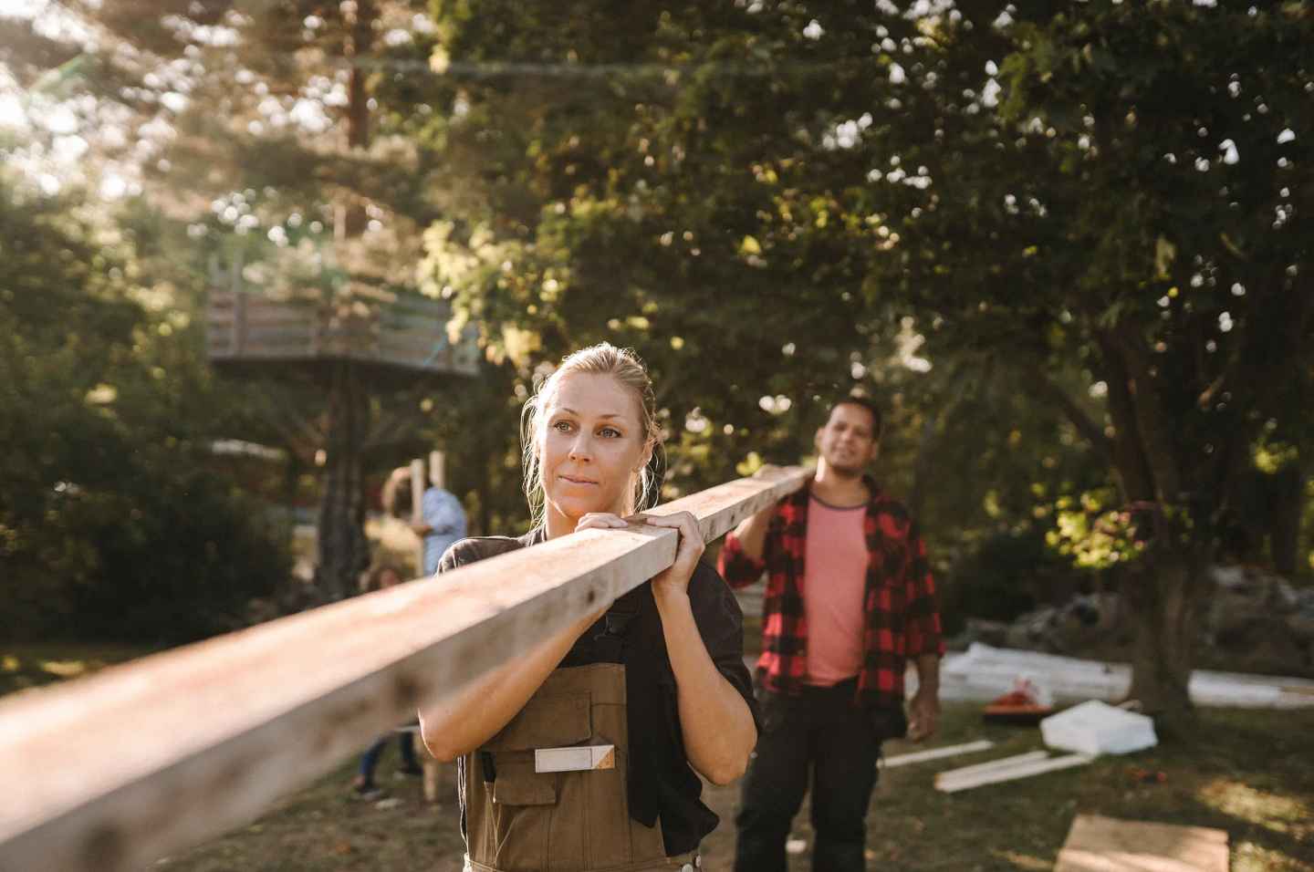 To tømrere bærer en lang planke sammen på en enkel byggeplass ute i skogen. 