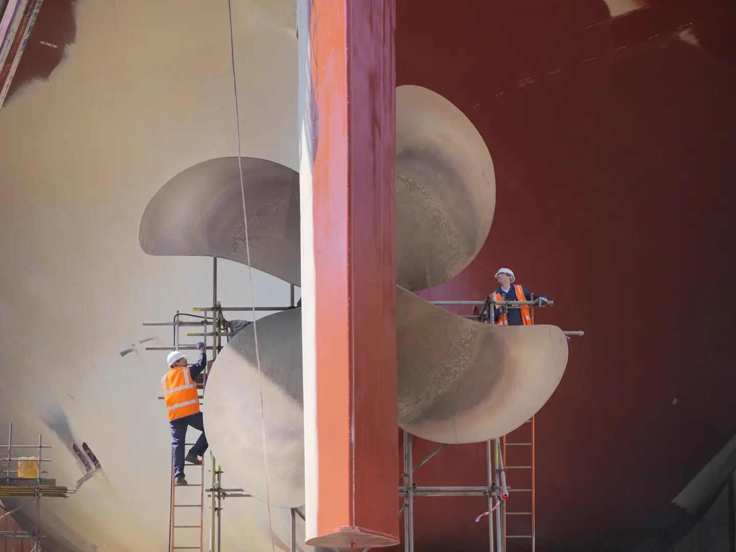 To arbeidere med hjelp og sikkerhetsvest vedlikeholder en stor båtpropell fra stiger