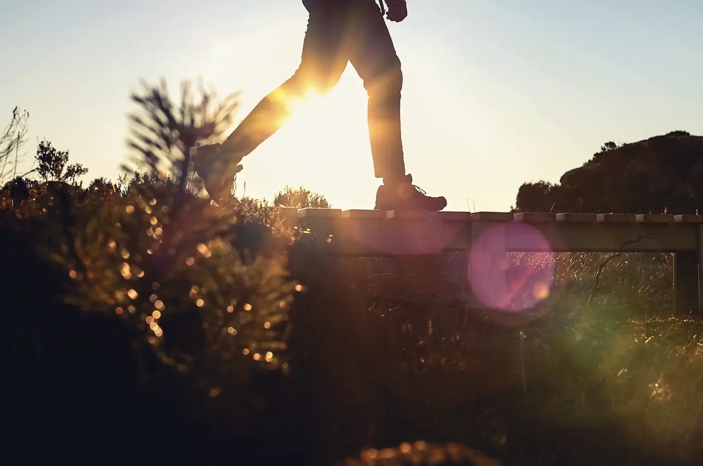 Person i fritidsklær går tur i åpen landskap, og krysser en bekk over en enkel trebro.