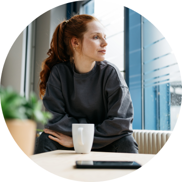 Kvinne sitter i en sofa og ser ut av vinduet. Grått vær, og kvinnen ser sliten ut.