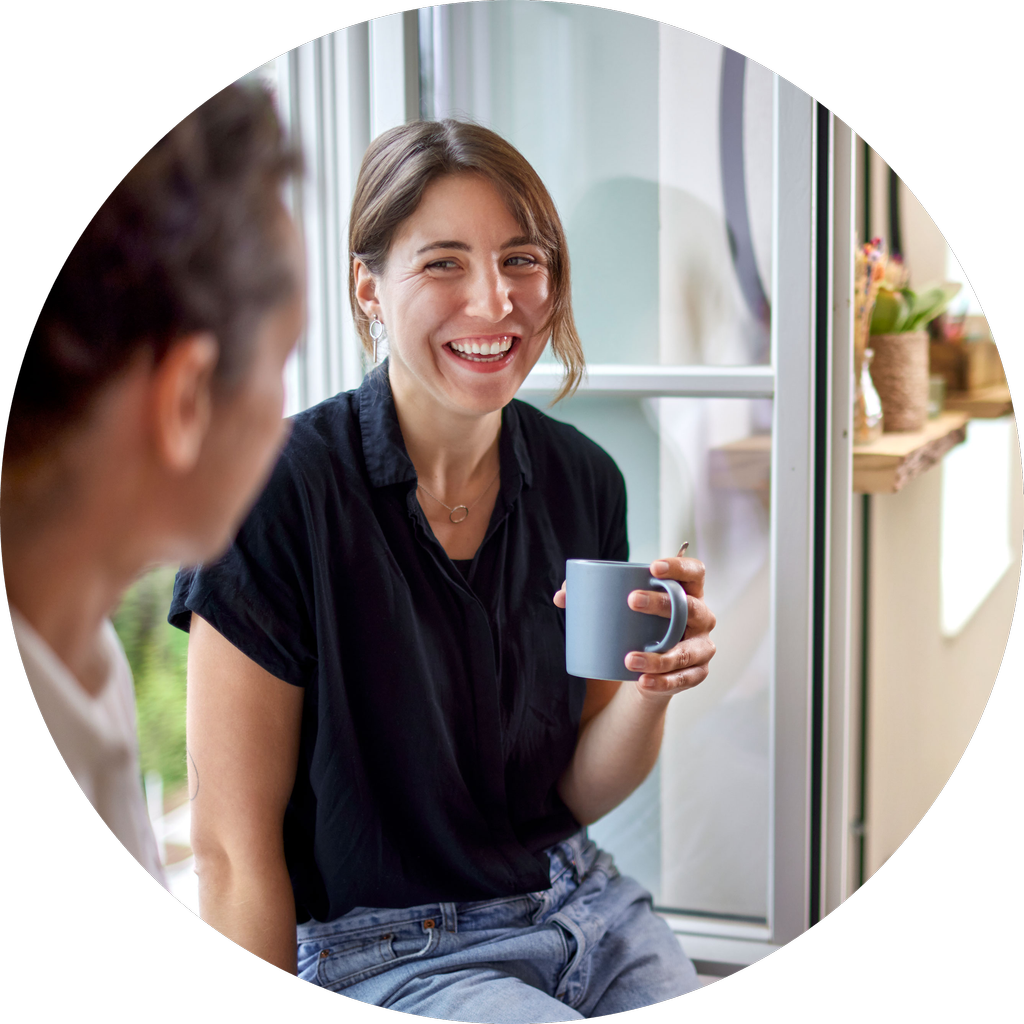 Person som sitter i vinduskarm med kaffekopp i hånda og ler til en kollega